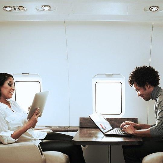 A man and a woman sitting at a table on a private jet working off a laptop and tablet with business aviation connectivity